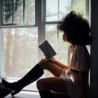 a girl sitting next to a window reading a book